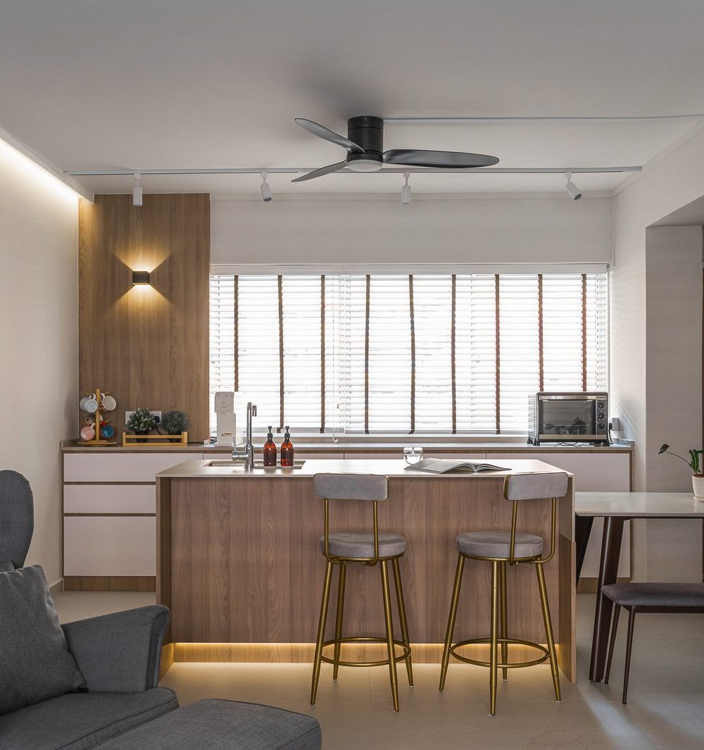 A timber-fronted island with brass-legged bar stools anchors the open kitchen, visible from a grey wingback armchair in the living area.