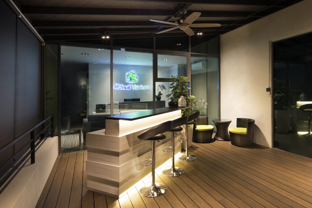 A backlit timber bar counter sits beneath a black slatted ceiling on the outdoor deck, paired with rattan lounge chairs finished in yellow cushions.
