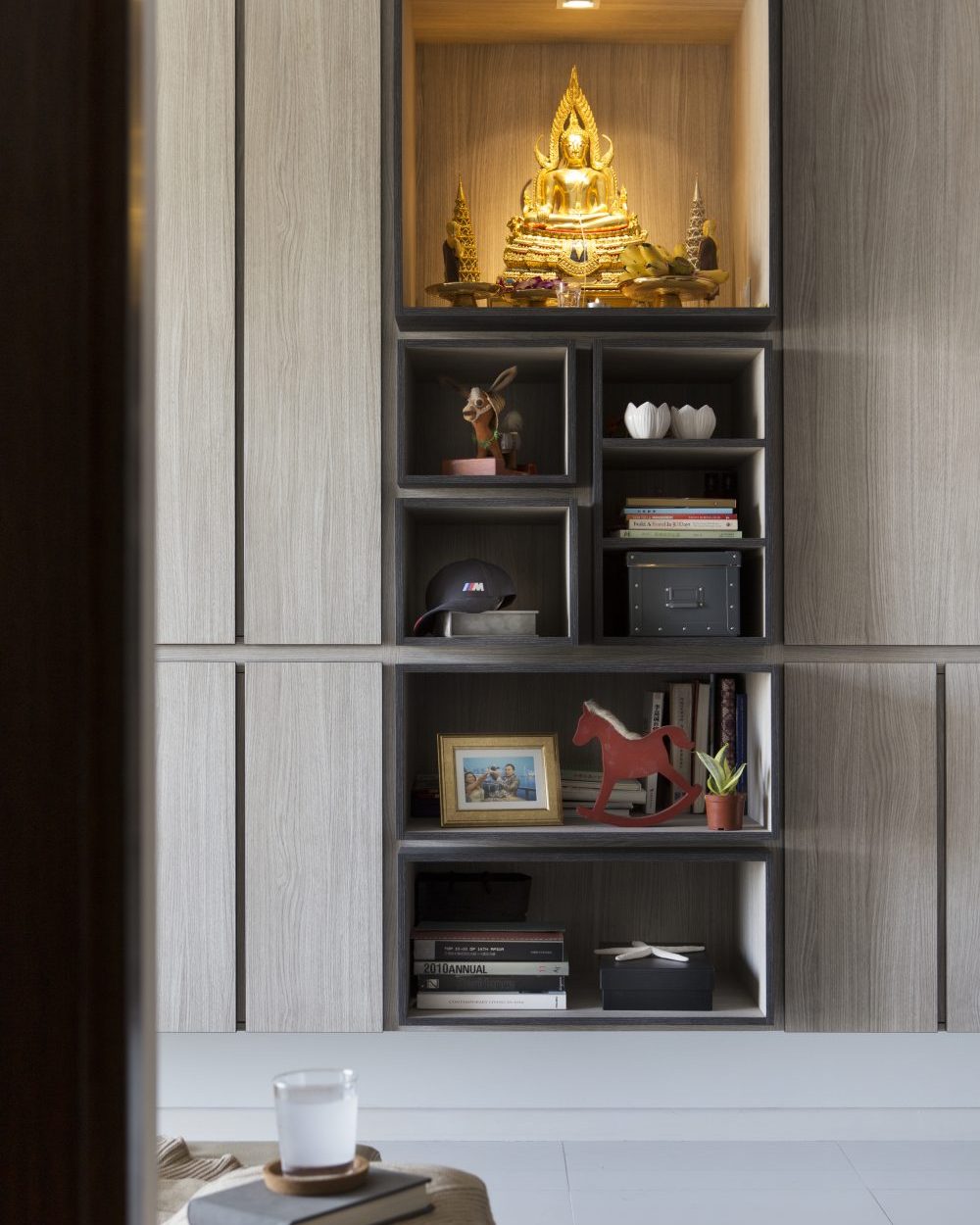 Open display boxes hold keepsakes, books and a vintage rocking horse, framed beneath the illuminated Buddha altar.