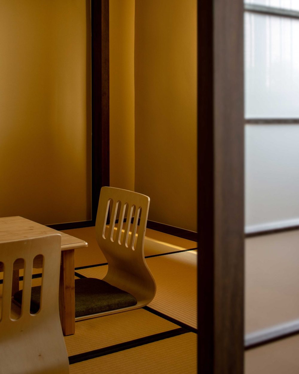 A single legless chair sits at the low tea table, set against warm mustard-yellow walls and tatami flooring.