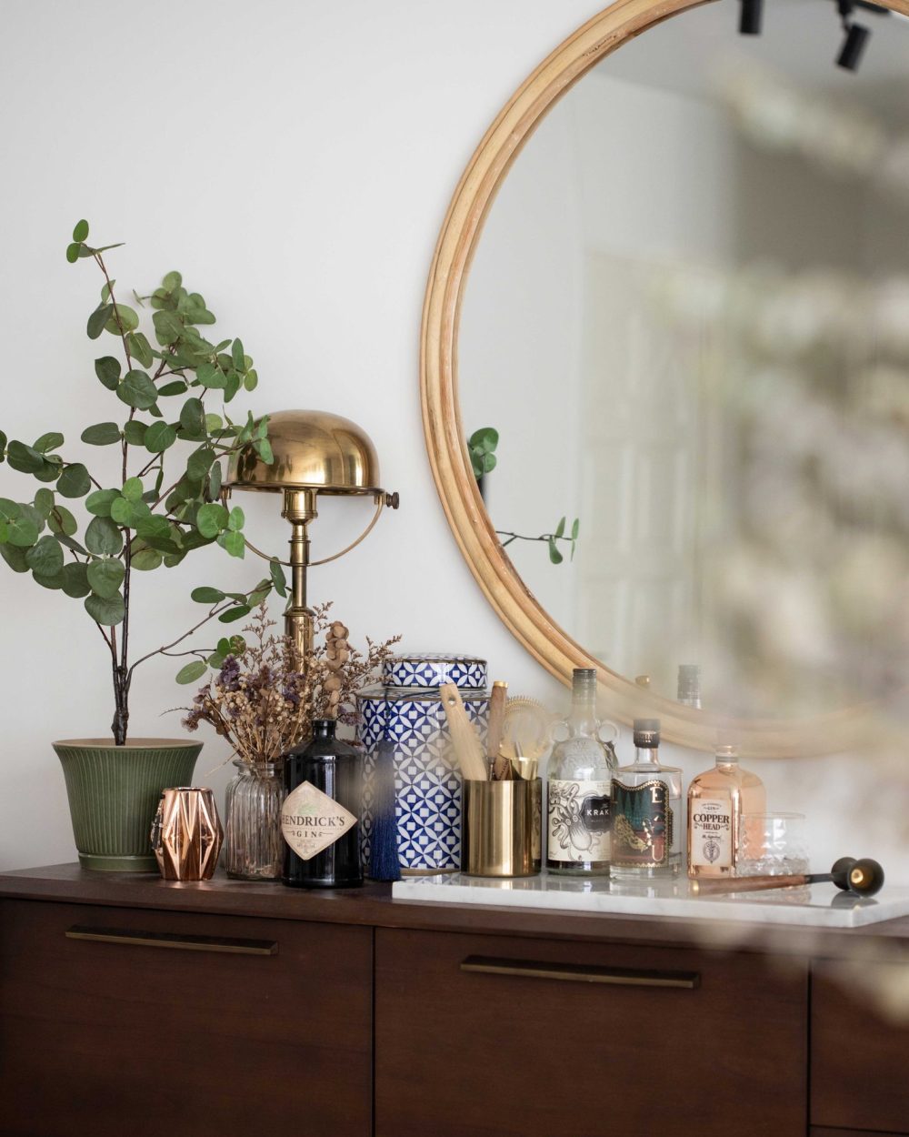 A rattan-framed round mirror hangs above a curated bar cart of gins and spirits, set on a walnut sideboard.