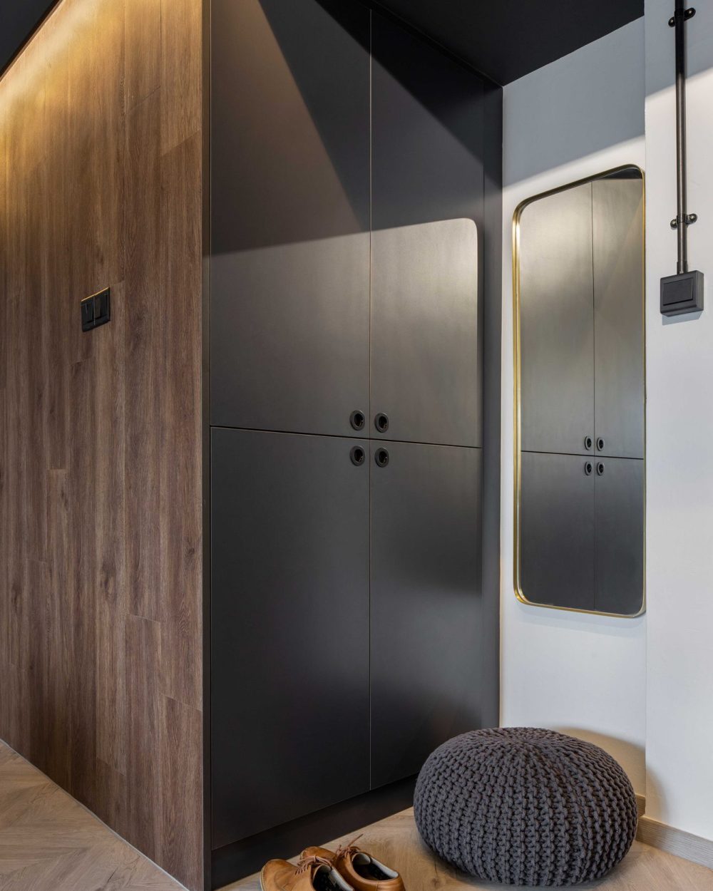 A matte black shoe cabinet with circular pulls sits beneath a brass-framed mirror, paired with a knit floor pouf.