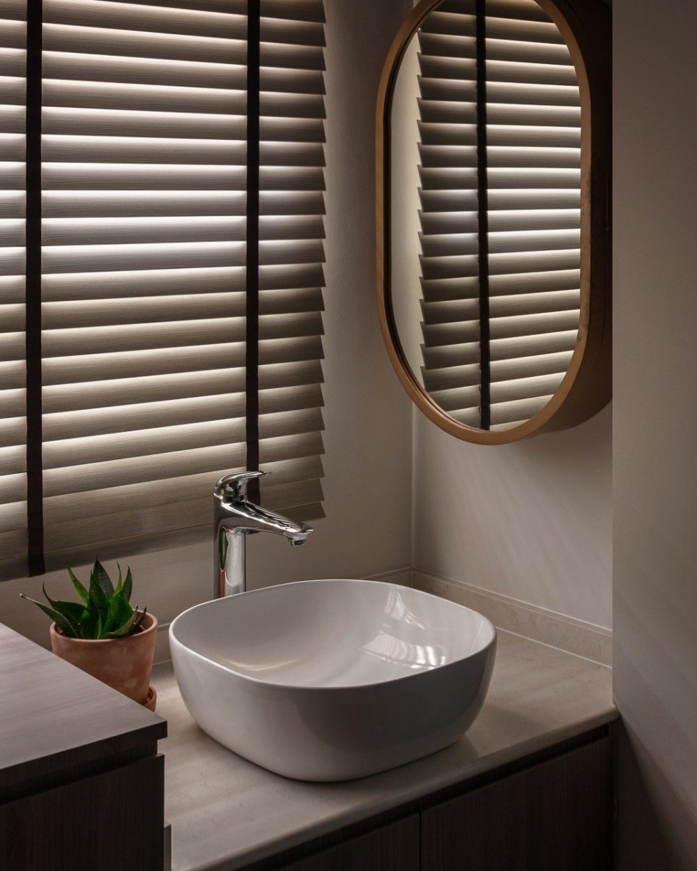 A rounded white vessel basin sits on a stone counter beneath an oval wood-framed mirror, with warm blinds and a potted succulent softening the corner.