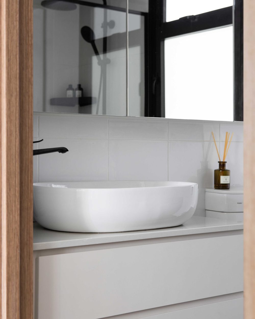 A sculptural white vessel sink and matte black tapware sit beneath a mirrored cabinet, reflecting the glass-enclosed rain shower behind.