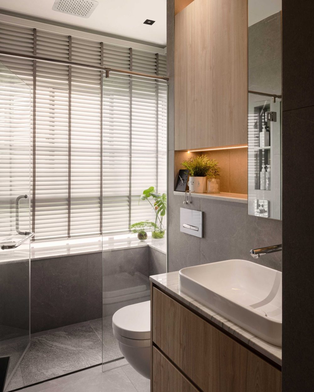 A rainfall shower and wall-hung toilet sit within grey stone tiling, softened by a built-in timber cabinet and potted greenery.