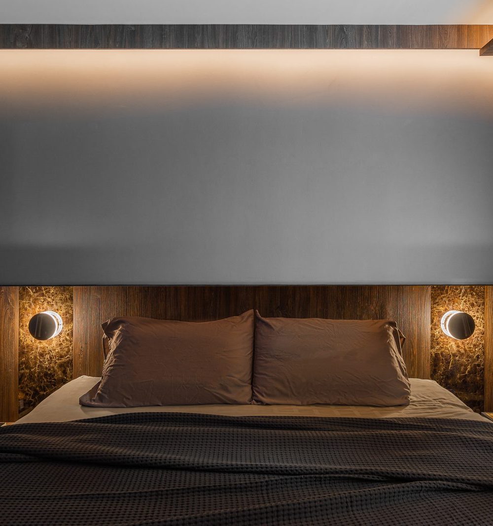 Warm wood-look panelling and a pair of dark marble insets with sconce lighting frame the bed, softened by cove lighting above.