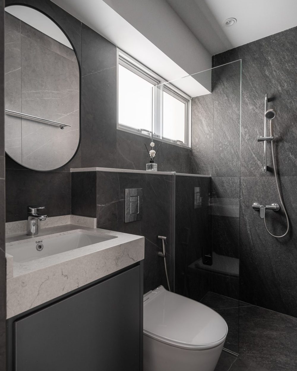 Dark slate-effect tiling wraps this bathroom's walls and floor, paired with an oval mirror and a glass shower divider.