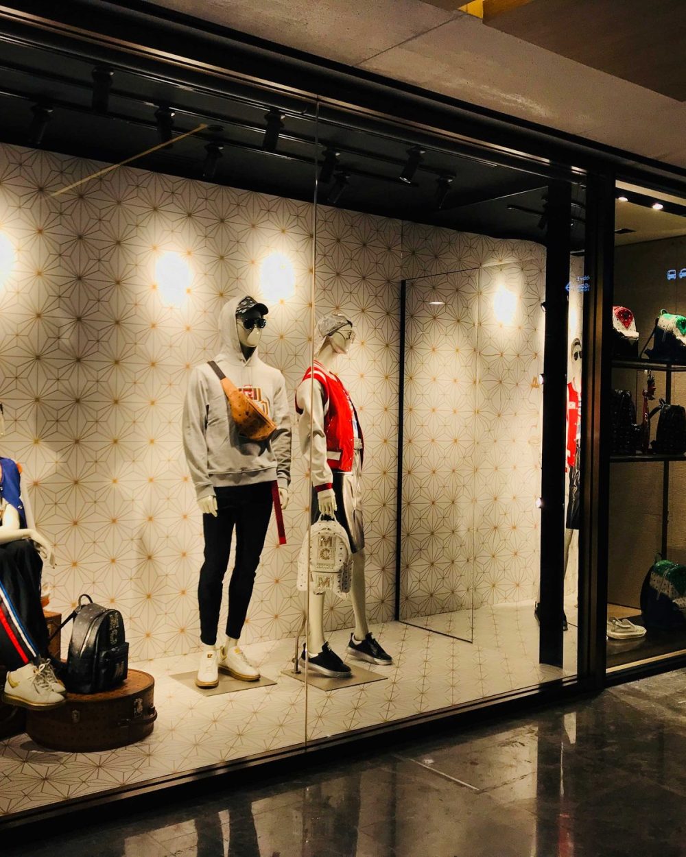 Three mannequins styled in outerwear in the MCM shopfront window against a geometric patterned wall.