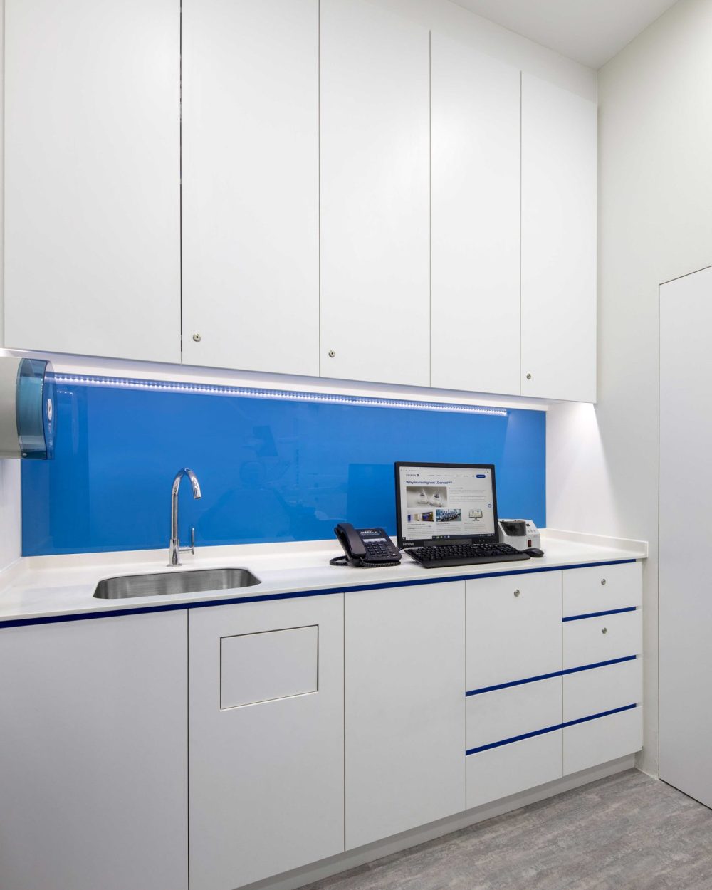 Glossy white cabinetry and a stainless steel sink keep the treatment room's wet area clean and efficient, framed by the same blue glass panel.