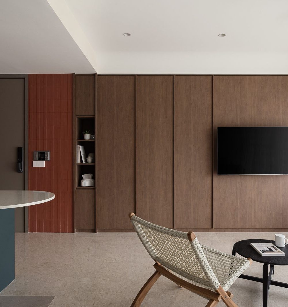 A terracotta tile wall and dark timber TV panel frame the entrance, with a teal kitchen island and woven bistro chair visible just beyond.