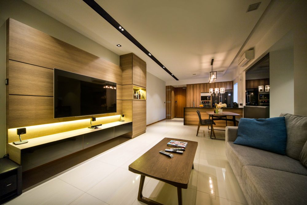A wide timber panel wraps the TV wall with backlit open shelving below, while industrial cage pendants mark the dining table beyond.