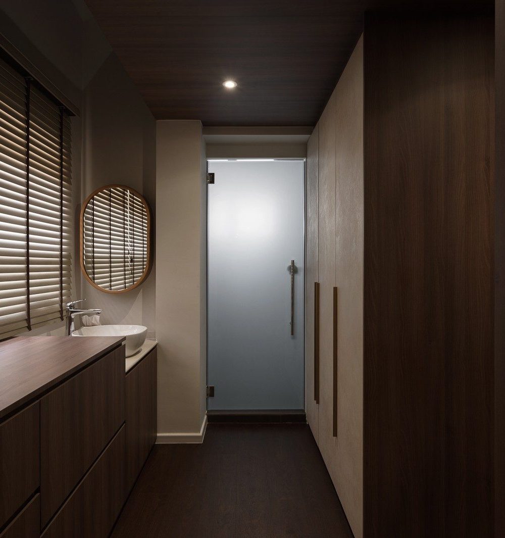 A run of dark timber wardrobes lines the walk-in corridor, with a wood-topped vanity and round mirror beneath the window and a frosted glass door lead