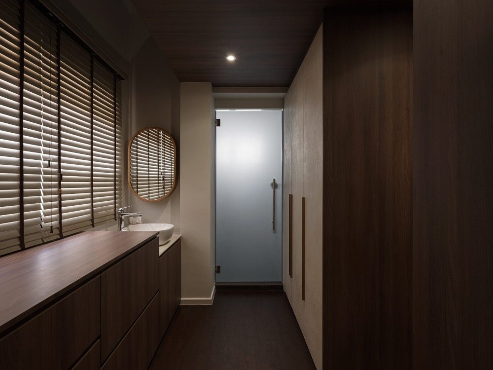 A run of dark timber wardrobes lines the walk-in corridor, with a wood-topped vanity and round mirror beneath the window and a frosted glass door lead