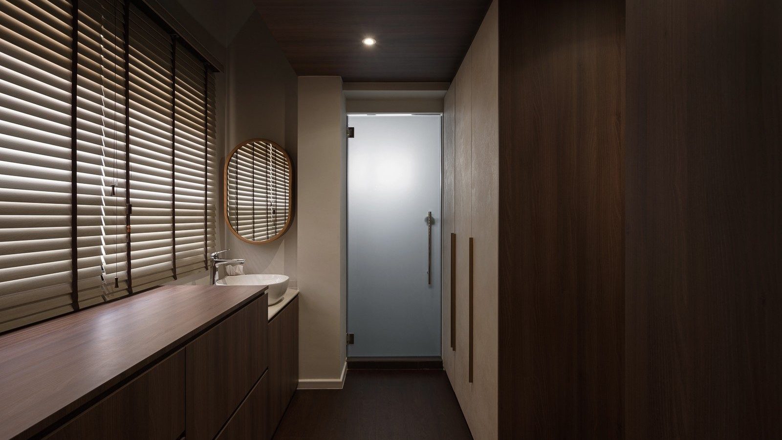 A run of dark timber wardrobes lines the walk-in corridor, with a wood-topped vanity and round mirror beneath the window and a frosted glass door lead