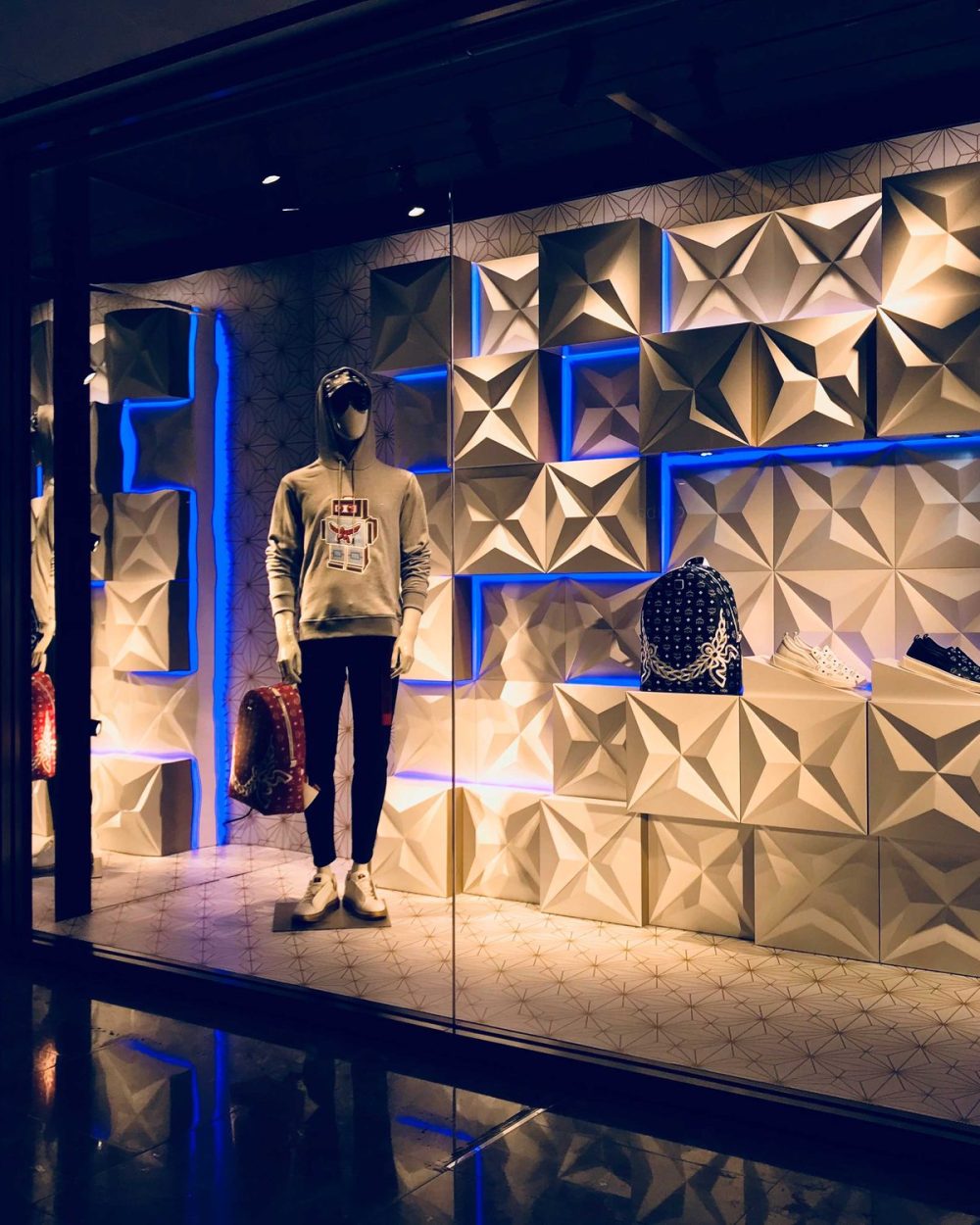 A hooded mannequin in MCM streetwear against a geometric matte white panel wall, holding a backpack.