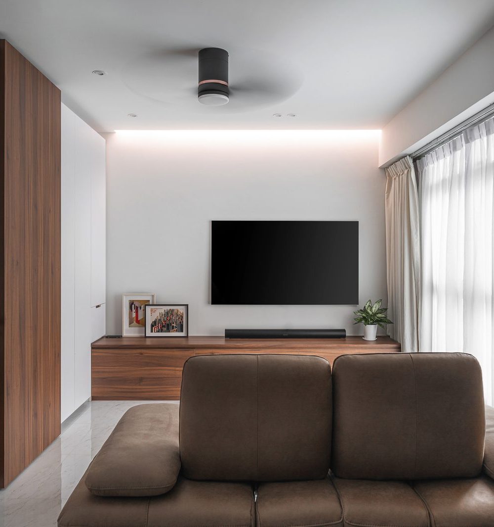 Walnut-toned cabinetry and a glass-fronted display cabinet frame the TV wall, warmed by a cove-lit ceiling and sheer curtains.