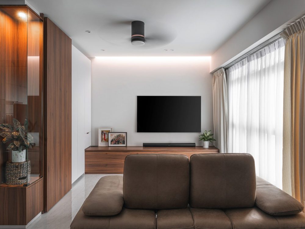 Walnut-toned cabinetry and a glass-fronted display cabinet frame the TV wall, warmed by a cove-lit ceiling and sheer curtains.