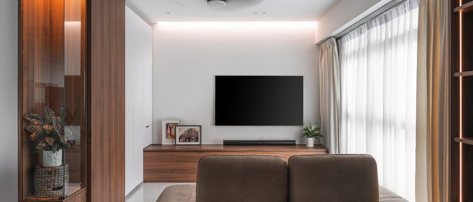 Walnut-toned cabinetry and a glass-fronted display cabinet frame the TV wall, warmed by a cove-lit ceiling and sheer curtains.