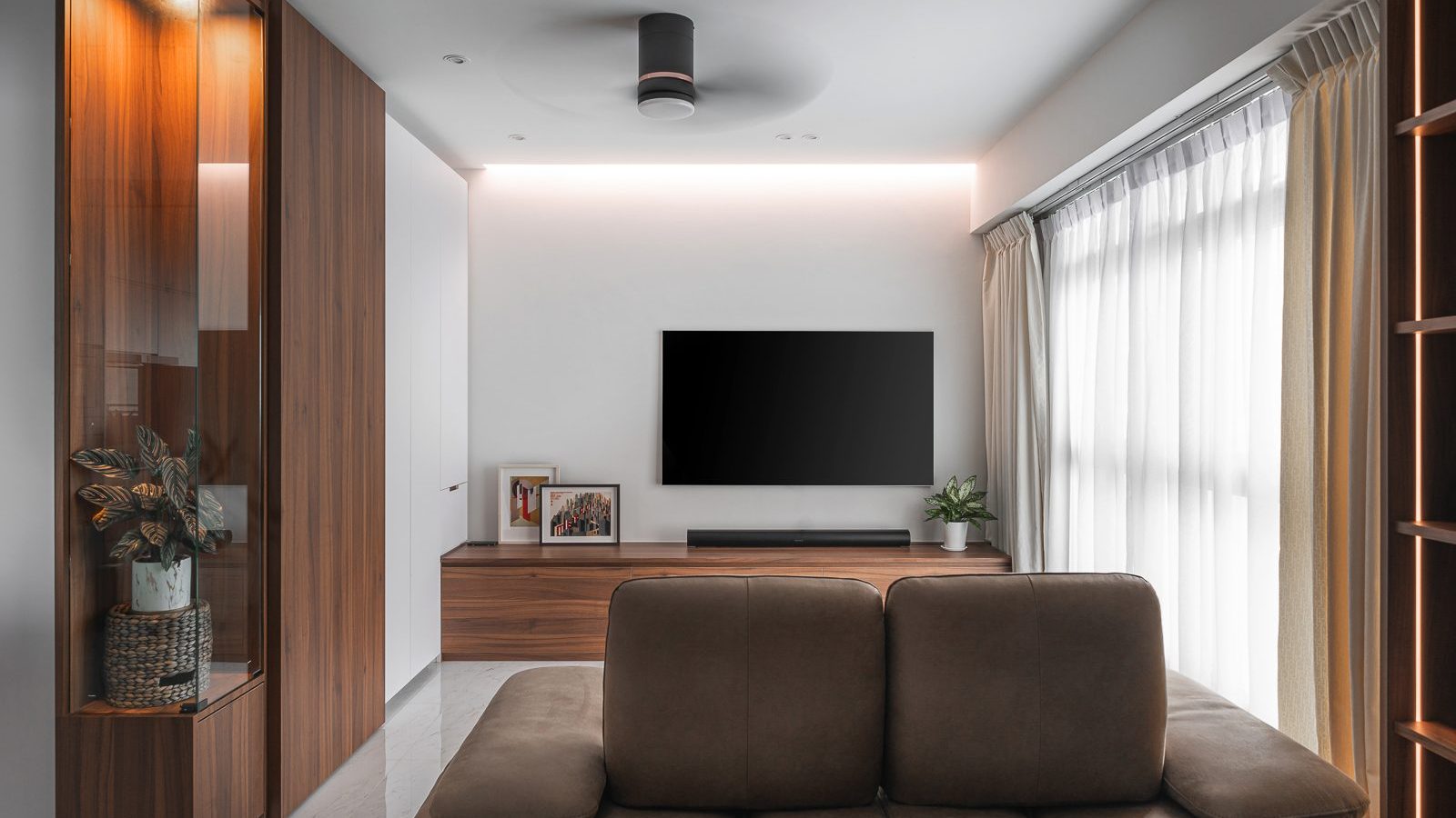 Walnut-toned cabinetry and a glass-fronted display cabinet frame the TV wall, warmed by a cove-lit ceiling and sheer curtains.