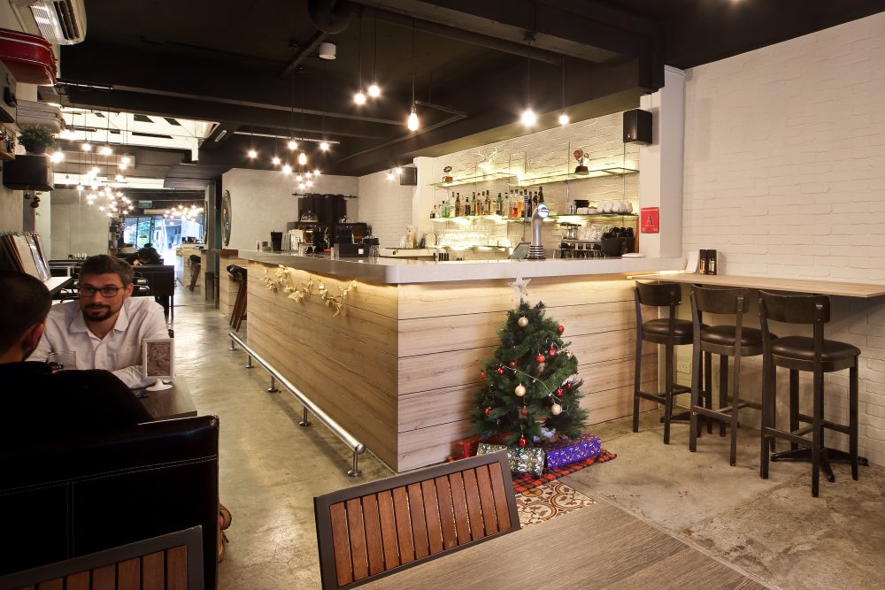 Exposed black ceiling beams and string pendant lights hang over a whitewashed brick bar counter, finished with glass shelving and a well-stocked back