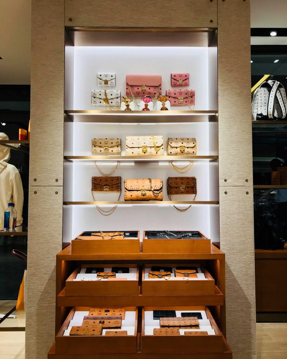 Wallets and chain-strap purses in pink, cream and tan displayed on illuminated shelving.