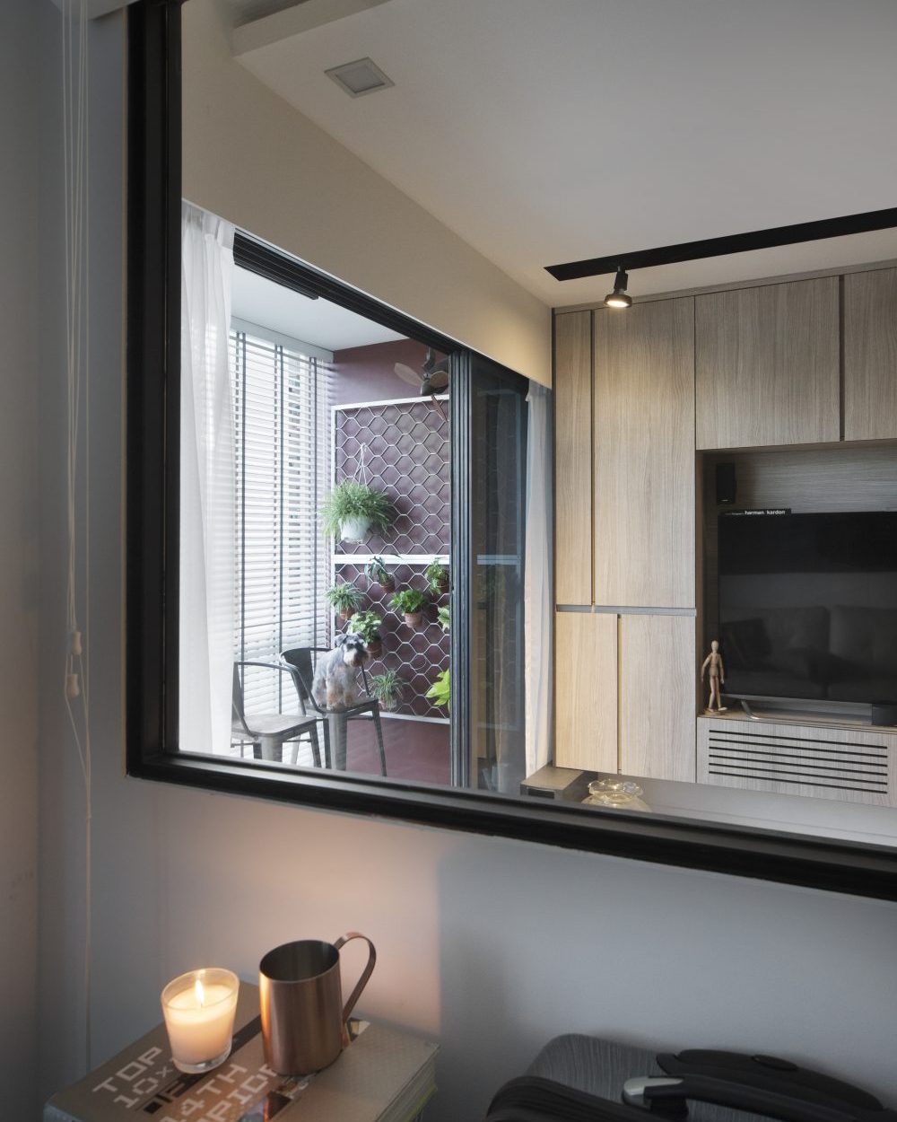 An internal window between bedroom and living area lets light and sightlines pass through, framing a view straight to the TV wall and balcony beyond.