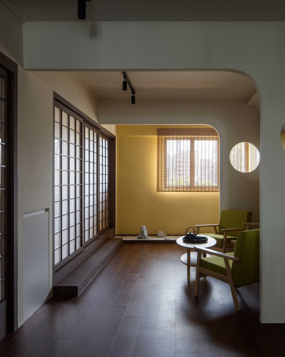 Olive green armchairs and a low round table sit beside a miniature rock garden, lit by a bamboo blind window.